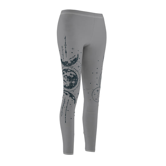 Front view of gray leggings featuring a vertical hand-drawn illustration of moon phases and geometric arrows on the left leg. The fabric has a soft, brushed suede texture with visible white stitching at the waist and hem.